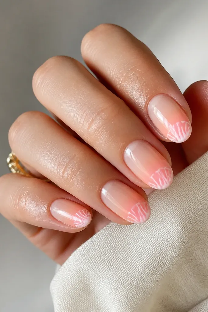 Short almond nails show a pastel ombre from sheer nude near the cuticle to soft peach and pale pink near the tips. On the tips, there are small milky white shell arcs, like tiny half-ribs.