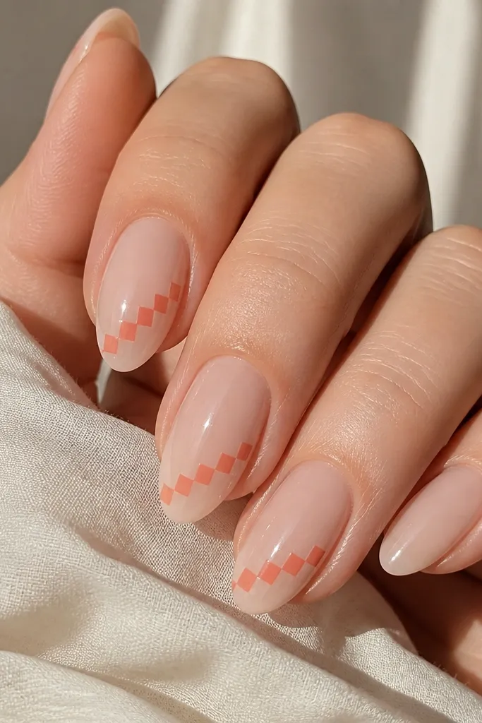 Short almond nails with a sheer nude base; three nails have a thin coral checkered strip running diagonally near the tip made of small squares; one nail is plain nude; glossy.