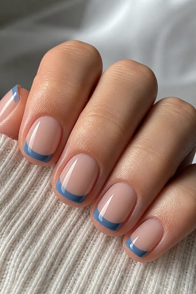 Five short square nails. Each nail has a nude pink base. The French tip is a medium denim blue with a clean curved smile line, glossy top coat, no extra art.