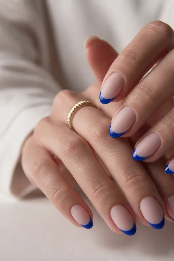 Short almond nails. Sheer nude base. Cobalt blue French tips that are glossy. The rest of the nail is finished with a matte top coat, creating a glossy-blue/matte-nude contrast.