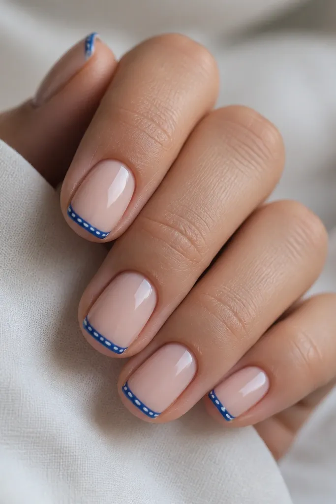 Short squoval nails with a nude base. Blue French tips. On each tip, there are 3-4 tiny white dashes spaced like small stitches, glossy finish.