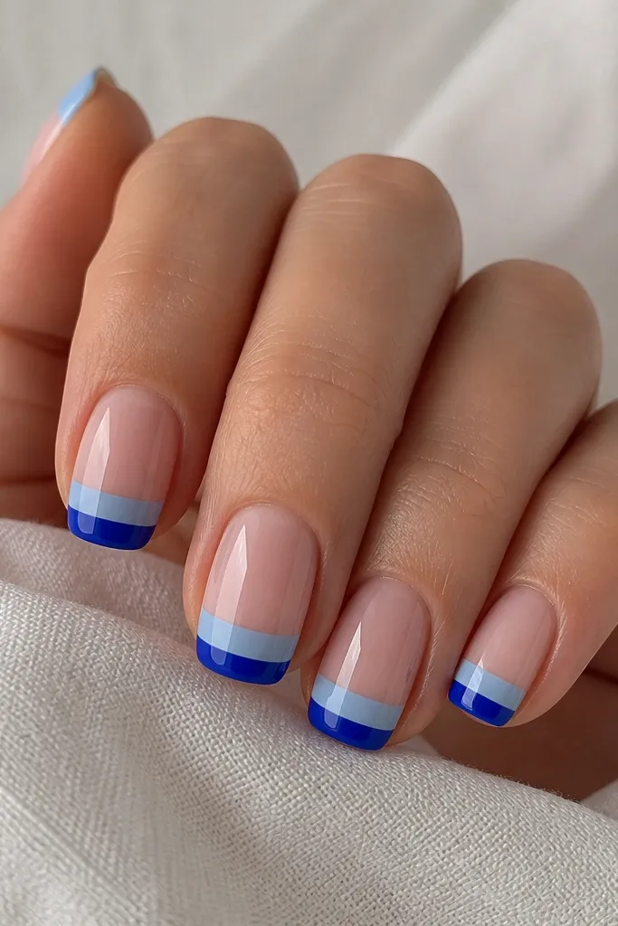 Short squoval nails. Nude base. Each French tip is split horizontally: top part baby blue, bottom part cobalt blue, with a sharp line between colors. Glossy finish.