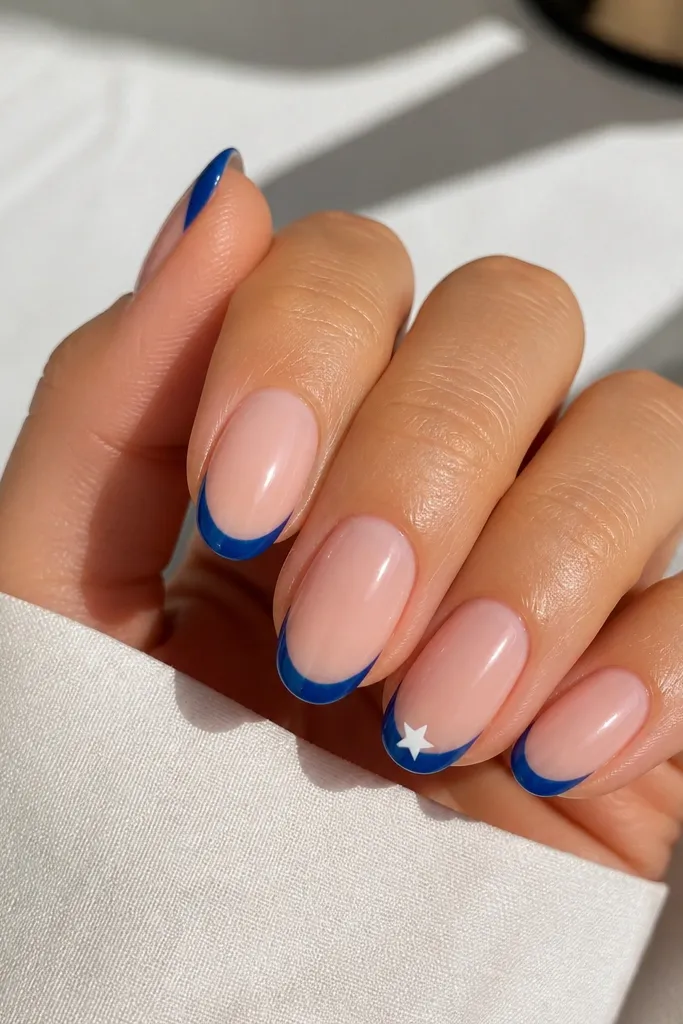 Short almond nails. Nude base. Blue French tips on all nails. On the ring finger, a single small white star sits on the blue tip near the center. Glossy finish.