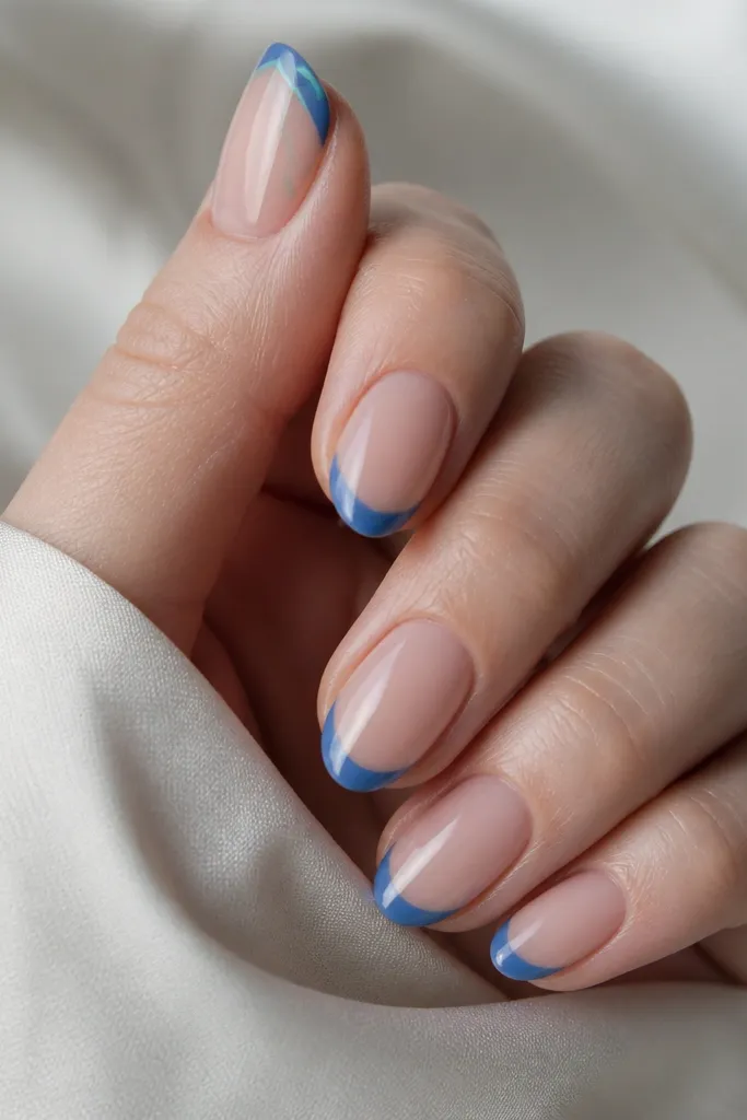Short almond nails. Nude base and blue French tips on all nails. On the thumb nail only, there's a small teal chevron drawn inside the blue tip area. Glossy finish.