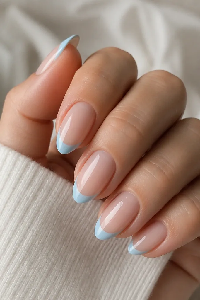 Medium almond nails with sheer nude base. A thin pastel sky blue French tip sits at the free edge. There's a negative space halo just below the tip at the cuticle side of the smile line, leaving a clear crescent.