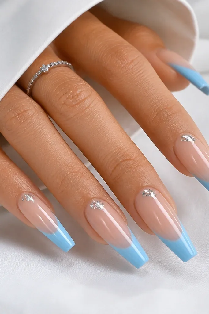 Long coffin nails with sheer nude base. Sky blue French tips are glossy and solid. At the cuticle corner of each nail, a small cluster of silver foil confetti sits near the side, not in the center.