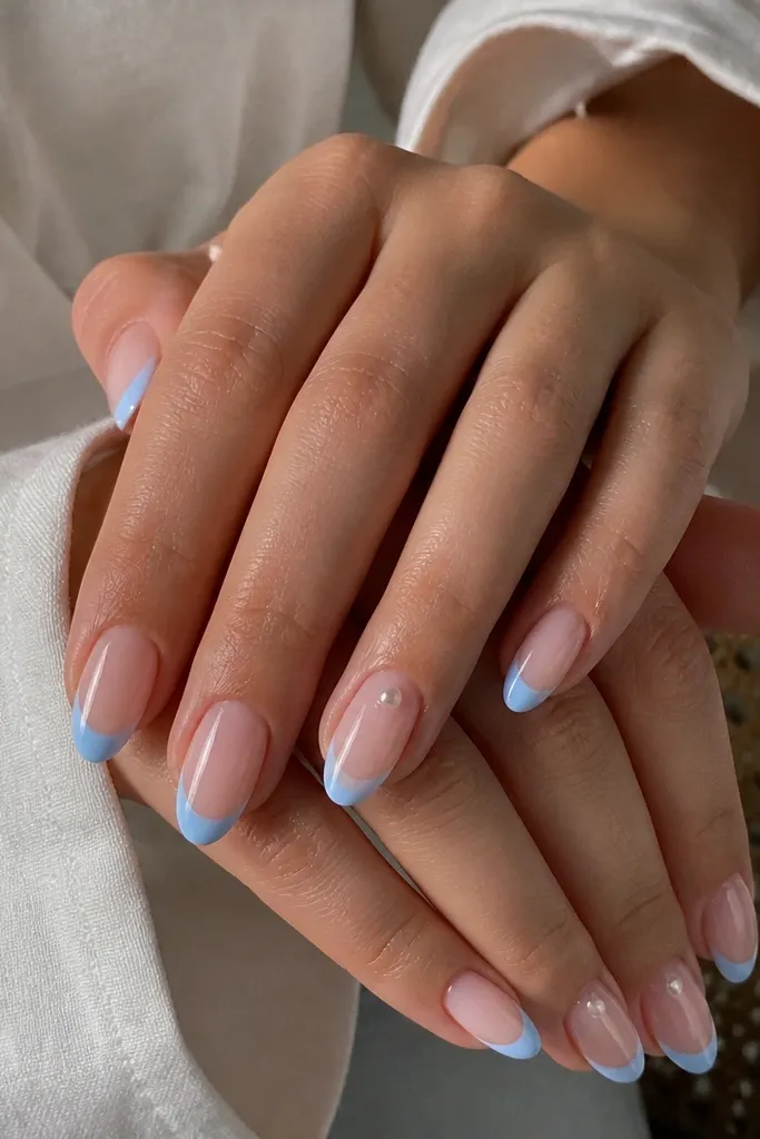 Medium oval nails with sheer nude base. French tips are sky blue, crisp and glossy. Ring fingers have a single small pearl dot near the cuticle on one side. Other fingers have no jewelry.