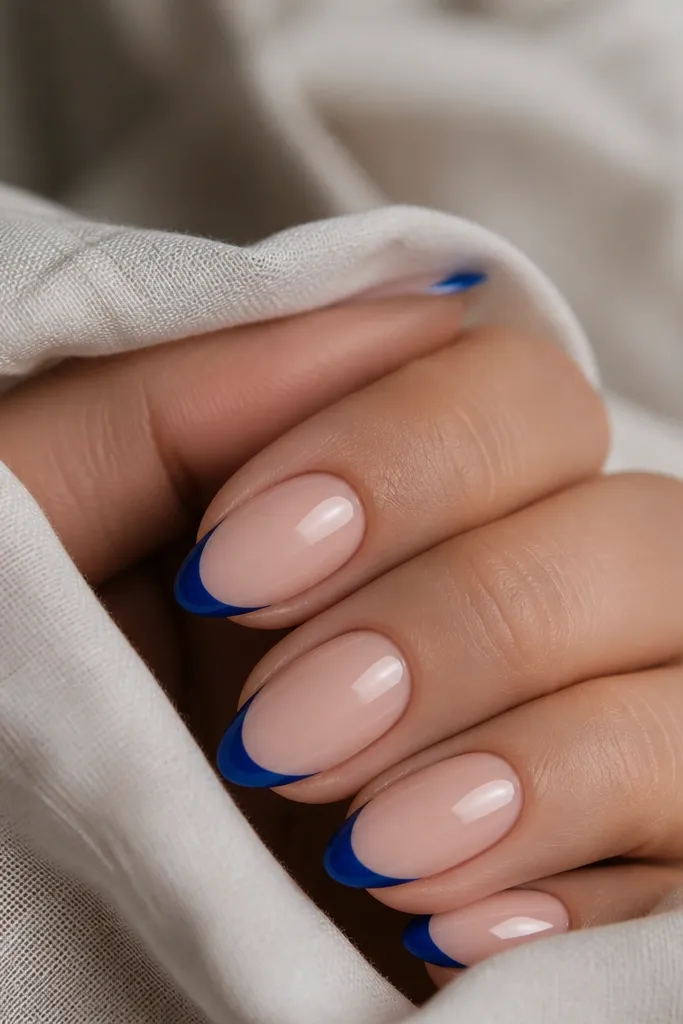 A close-up of short almond nails. The base is sheer nude-pink, and the tips are a tight cobalt-blue French line only about 2 mm wide at the center. The blue looks opaque and glossy, with a crisp curved edge and no bleeding into the nail bed.