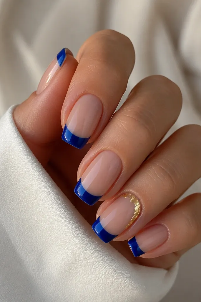 Nude base nails with royal blue French tips. The ring finger has a small strip of gold foil near the cuticle or along the side, while the other nails keep plain royal blue tips. Glossy top coat makes the blue look deep.