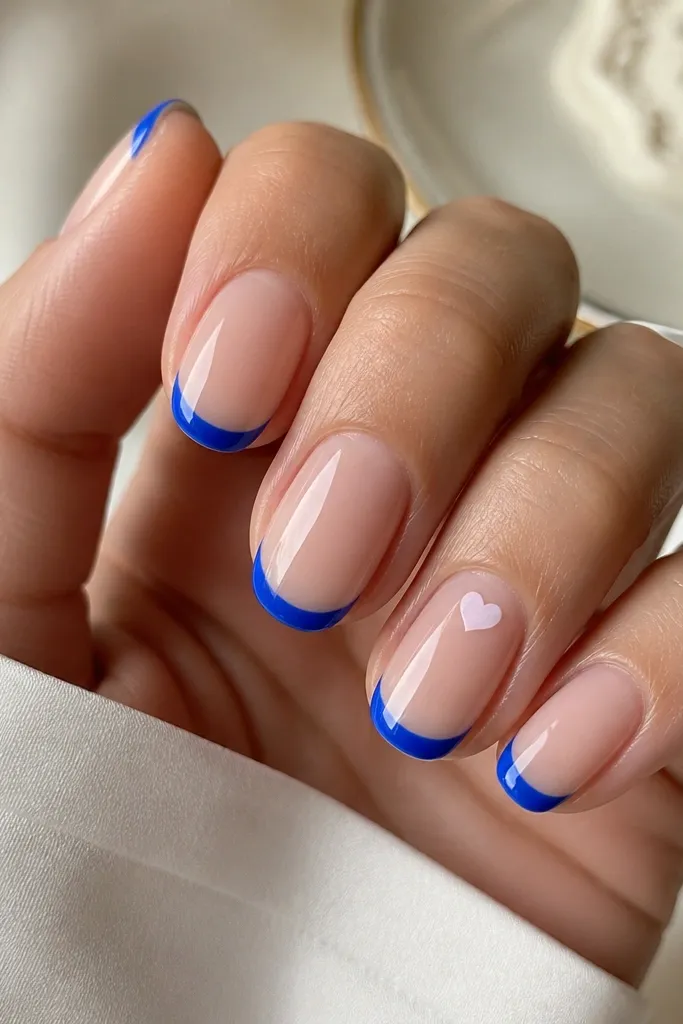 Nude base nails with cobalt-blue French tips on most fingers. One accent nail has a tiny micro heart drawn in white or pale blue near the cuticle area. Glossy top coat makes the heart look crisp.