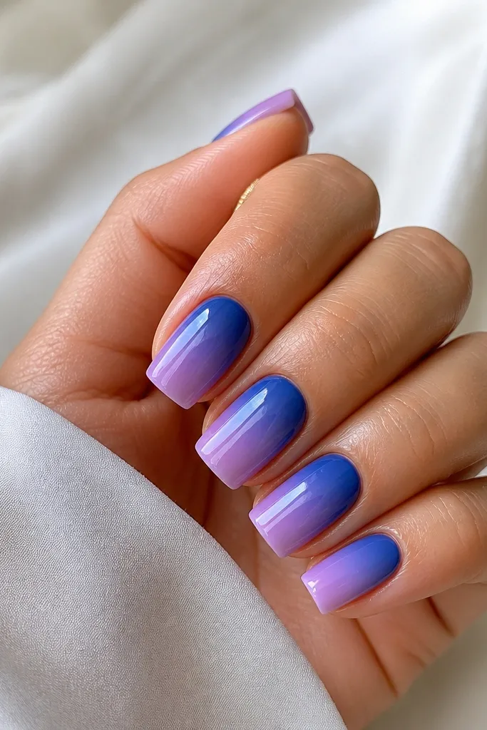 Square nails showing an ombré from deep blue at the cuticle fading into lilac at the tip. The finish is glossy but still slightly translucent, like a jelly glaze over the color.