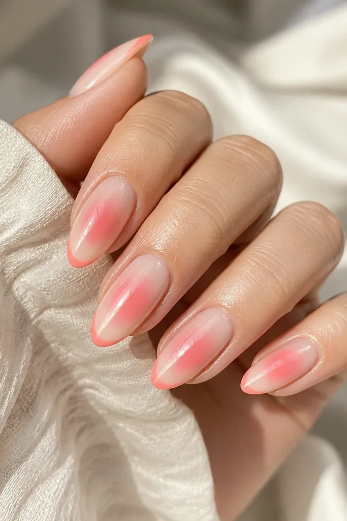 Almond nails with a soft pink aura glow around the center and a coral edge at the tip, surrounded by a milky nude base, glossy finish.