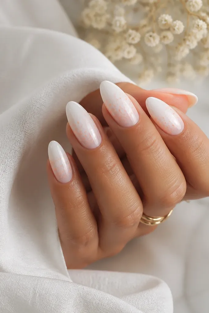 Almond nails with a milky white base; the tips have tiny peach sprinkle dots and flecks fading upward, glossy finish.