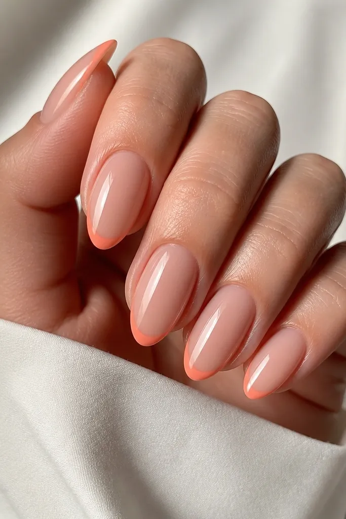 Almond nails with a nude base. The tips have a thin coral French line, crisp and close to the free edge. The rest of the nail is sheer and clean, with a high-gloss finish.