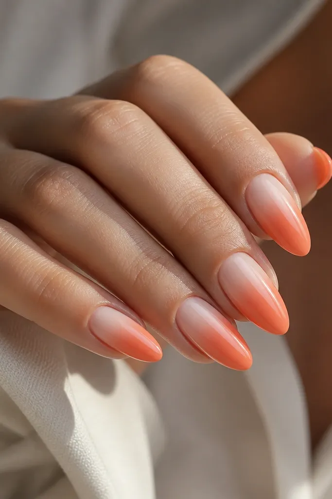 Almond nails with an ombre that starts sheer near the cuticle and blends into a coral-orange shade toward the tip. The gradient is smooth and glossy.