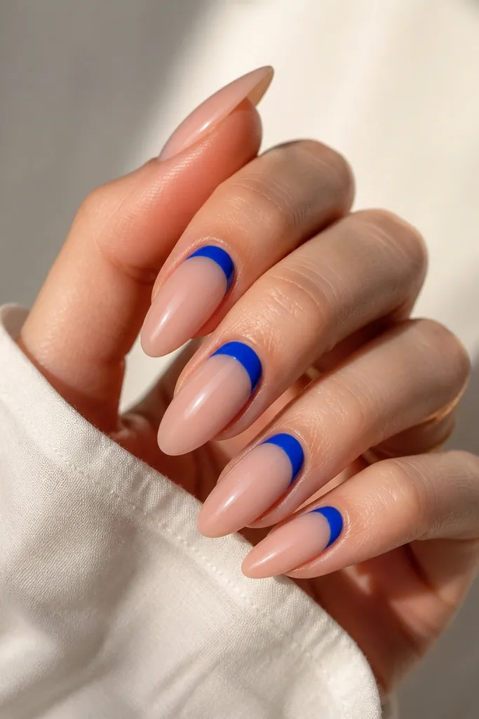 Almond nails with a nude base and a neon blue half-moon near the cuticle, leaving negative space around it. The blue shape is clean and glossy.