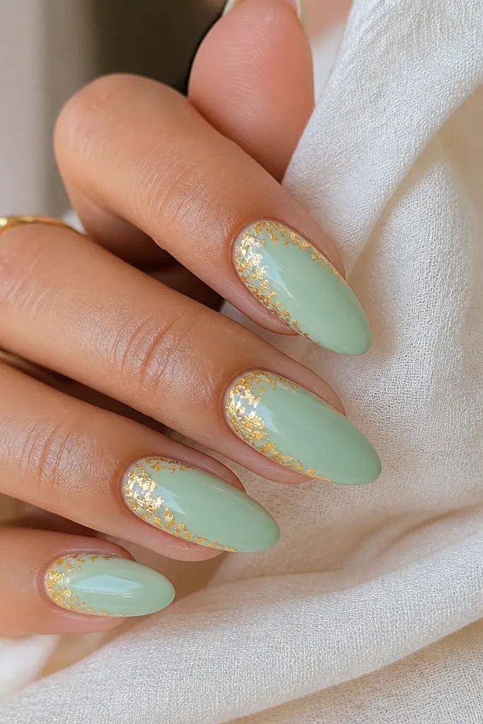Almond nails with mint base and irregular gold foil flakes concentrated near the cuticle and along one side; glossy.