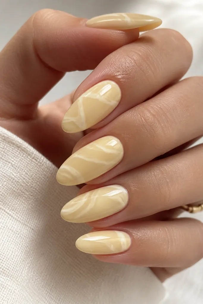 Almond nails in a creamy butter-yellow nude. Over the top, there are thin white marble swipes that look like soft streaks, not thick veins. The nails are glossy and smooth.