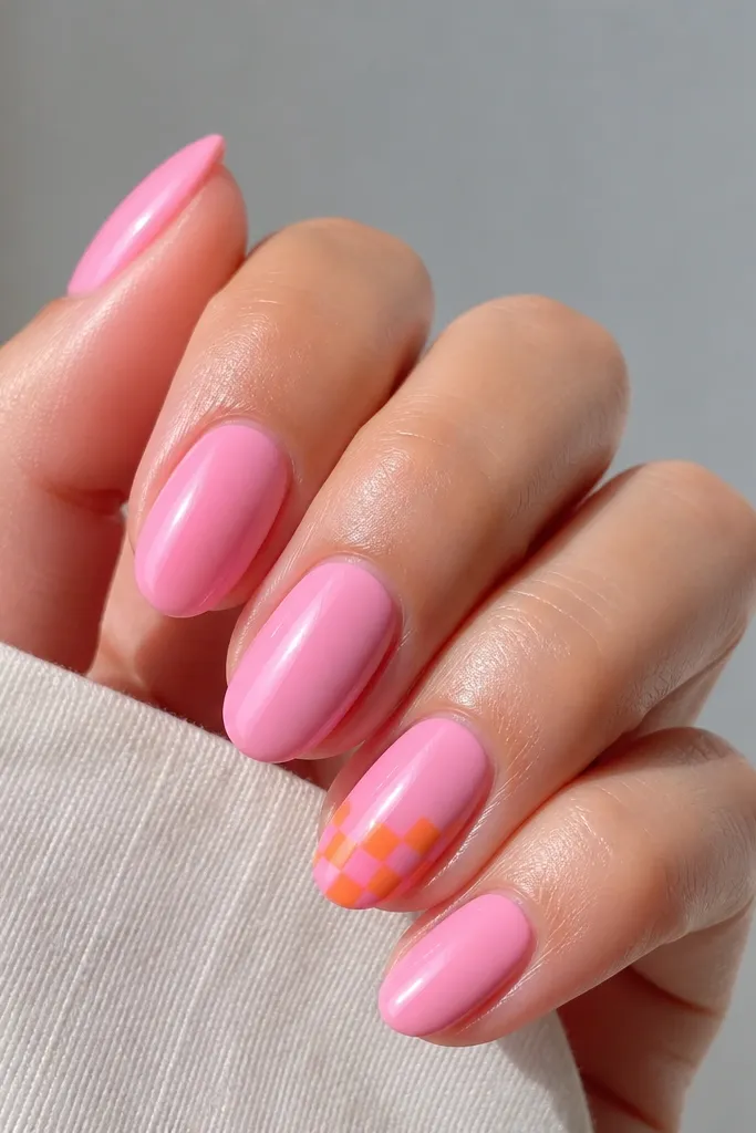 Pink nails with glossy finish. On one accent nail, there is a tiny checker pattern made of alternating orange and pink squares near the tip, with clean edges and a smooth top coat.