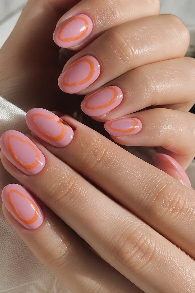 Oval nails painted glossy pink. Each nail has a thin orange swirl drawn in a loose circle around the center, like a halo ring, with negative space inside and outside the swirl.