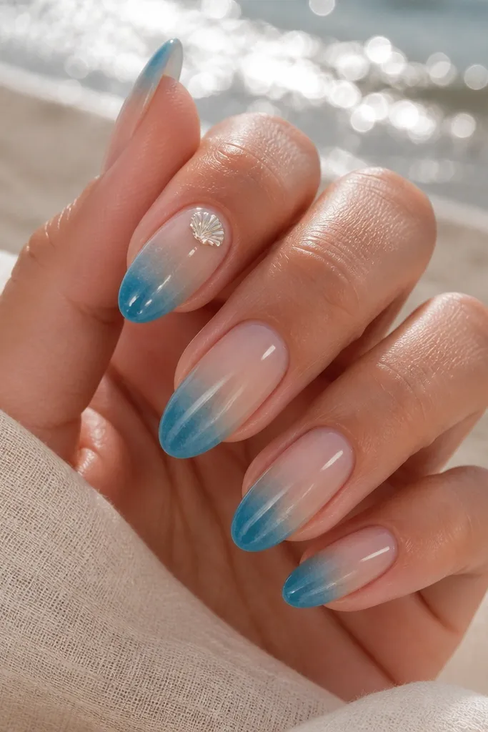 Medium almond nails with an ocean blue fade at the tips blending into sand nude at the base, one small shell charm sticker near the cuticle, glossy.