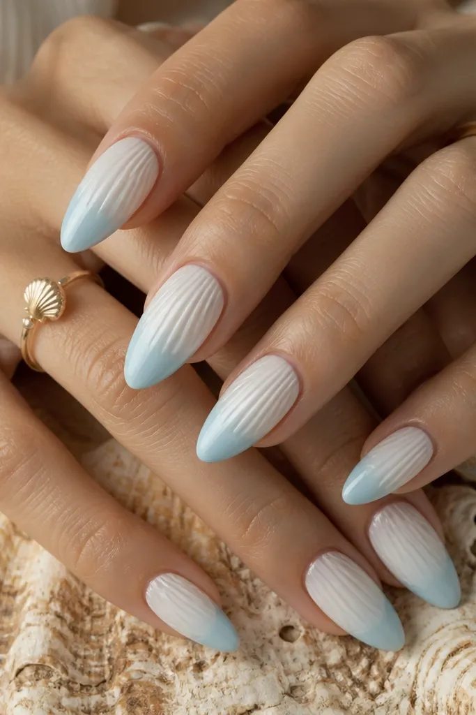 Long almond nails. The top half is milky white. The bottom third fades into a light blue-tinted milky ombré, like ocean at the shore. Shell ridges are painted mostly on the top half and continue subtly into the fade.